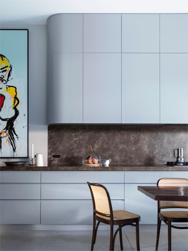 A refined modern kitchen and dining area with pale blue-grey flat-panel cabinetry, a dark marbled stone splashback and benchtop, integrated appliances, and a dark timber dining table with woven cane chairs. A curved cabinetry end and framed abstract artwork add sculptural detail.