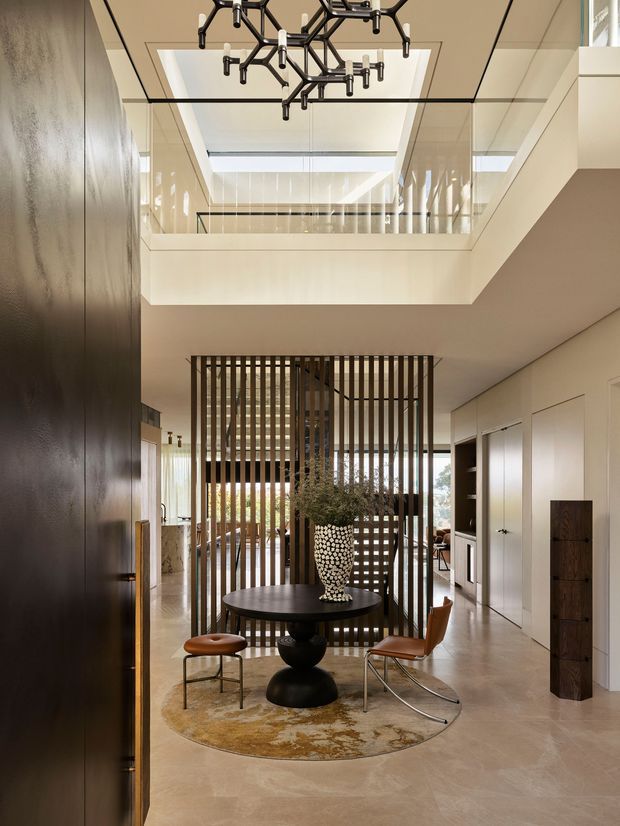 A refined contemporary interior foyer or entry hall with a double-height void and glass balustrade above. A sculptural black branching pendant hangs from the ceiling, while a dark timber vertical slatted screen separates the entry seating area from spaces beyond. The room features pale stone or polished concrete flooring, light neutral walls and cabinetry, dark timber wall paneling, a round black table, curved leather chairs, and a sculptural ceramic vase. The space is bright, spacious, and carefully composed with strong vertical and geometric elements.