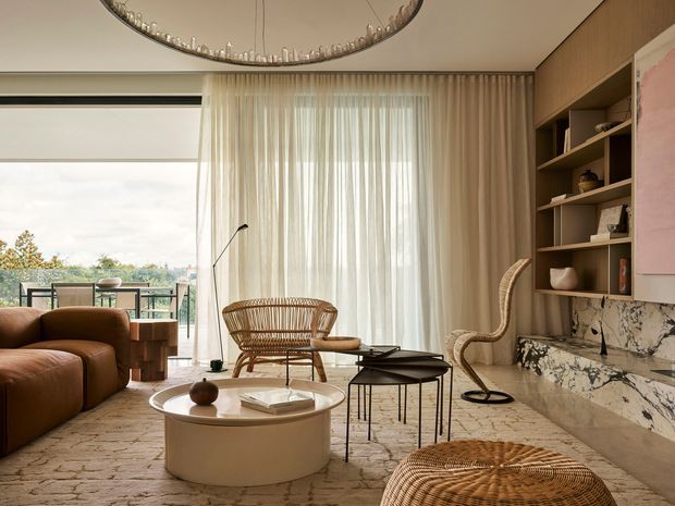 Warm, refined contemporary living room with a curved cream sofa, woven rattan lounge chairs and ottoman, round coffee table, layered neutral rug, and a dramatic marble-clad built-in feature wall with open shelving. Full-height sheer curtains frame glazing to a balcony and landscape beyond, while a sculptural circular pendant adds a decorative focal point.