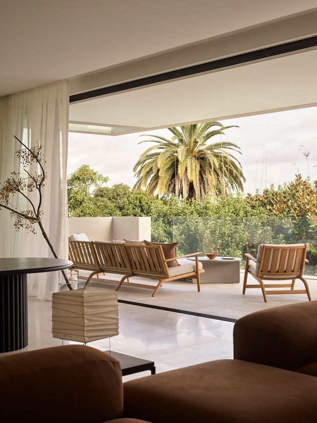 A refined contemporary indoor-outdoor living space opening onto a terrace. The room features floor-to-ceiling glazing, sheer curtains, polished light flooring, sculptural dark furniture, and a large sliding opening. Outside, a timber lounge setting sits behind a frameless glass balustrade, surrounded by dense greenery and a mature palm tree.
