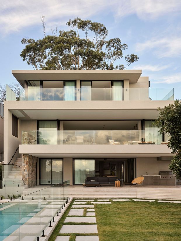 Photograph of a contemporary multi-storey residence with a flat roof, broad cantilevered horizontal slabs, extensive floor-to-ceiling glazing, and glass balcony balustrades. The pale rendered facade is complemented by sections of natural stone cladding. A landscaped lawn, stepping-stone path, terrace, and glass-fenced swimming pool occupy the foreground, while mature trees rise behind the house.