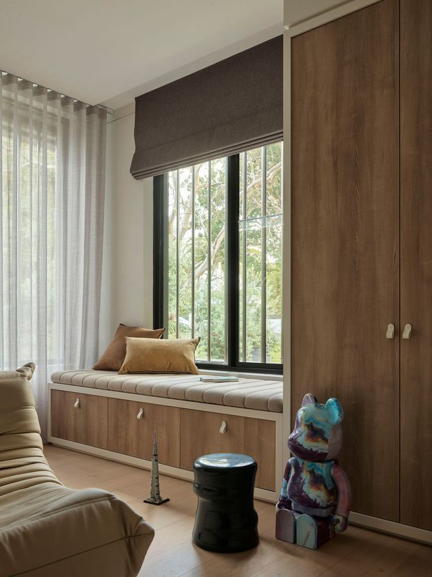 A refined living room corner with a built-in window seat beneath a tall, black-framed window, custom wood cabinetry, soft upholstered seating, and layered window treatments. The room features warm timber joinery, neutral upholstery, sculptural side tables, and a colourful decorative bear sculpture, with leafy greenery visible outside.