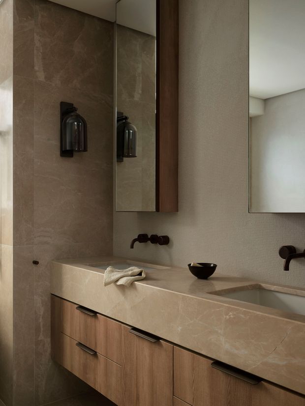 A refined contemporary bathroom vanity with a long stone countertop, integrated basins, warm wood cabinetry, large mirrors, textured wall finish, and dark sculptural wall-mounted tapware and lighting. The space has a warm, muted neutral palette with layered stone and timber finishes.