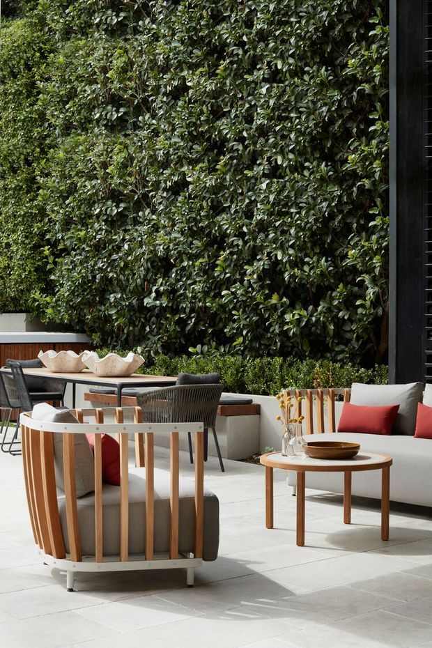 A refined outdoor lounge and dining terrace with pale stone paving, contemporary timber and woven furniture, a low coffee table, and a large dense green living wall providing a lush backdrop. Neutral upholstery is accented with red cushions and natural timber tones.