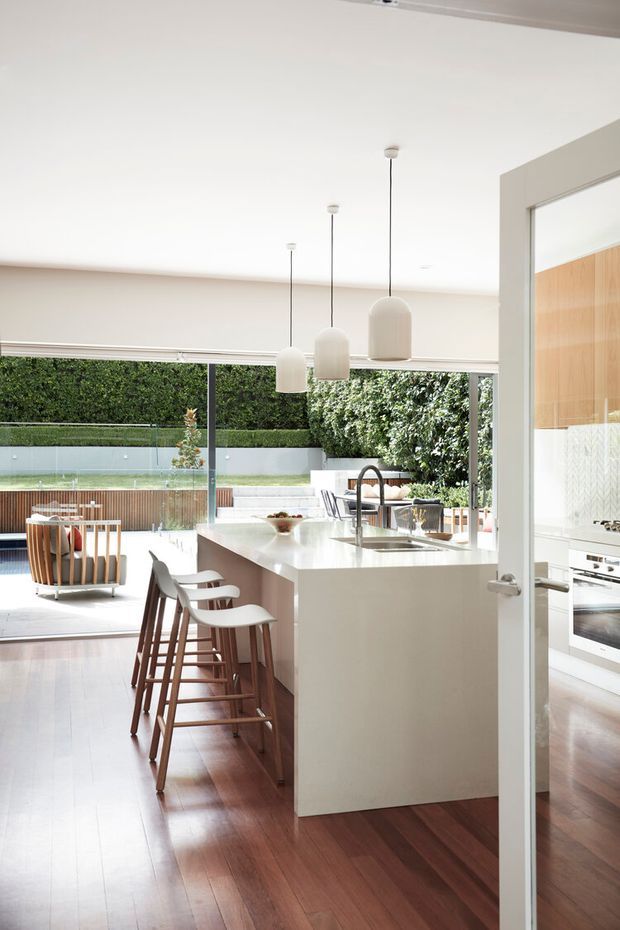Bright contemporary kitchen with a large white island, integrated sink and gooseneck tap, timber bar stools, white pendant lights, and warm timber flooring. Full-height glazing opens onto a landscaped courtyard with a swimming pool, while a secondary kitchen run is visible through the doorway.