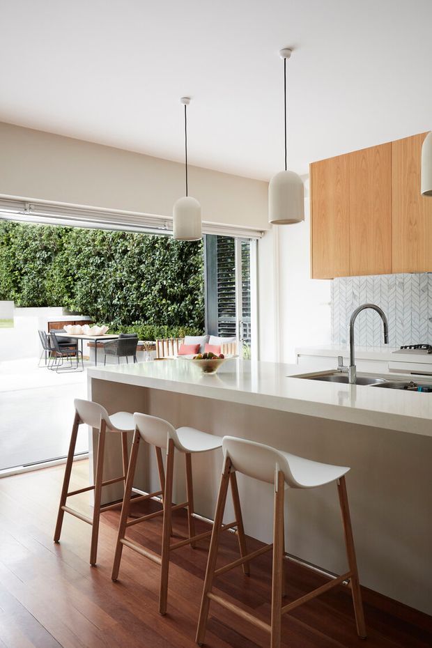 Bright contemporary kitchen with a large white island and integrated sink, four timber-framed bar stools, pale timber cabinetry, and a patterned light backsplash. Pendant lights hang above the island, while full-height glazing and a lush green wall provide an indoor-outdoor connection to the dining area and courtyard beyond.
