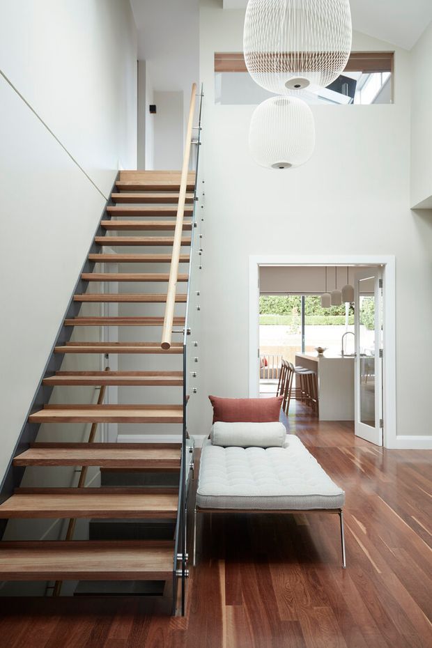 Bright contemporary interior featuring an open-riser timber staircase with dark metal supports and a glass balustrade, leading into a double-height living area. Two oversized white pendant lights hang overhead, while a white upholstered chaise with a rust-coloured cushion sits on polished timber flooring. A glazed opening reveals a dining or kitchen area beyond.