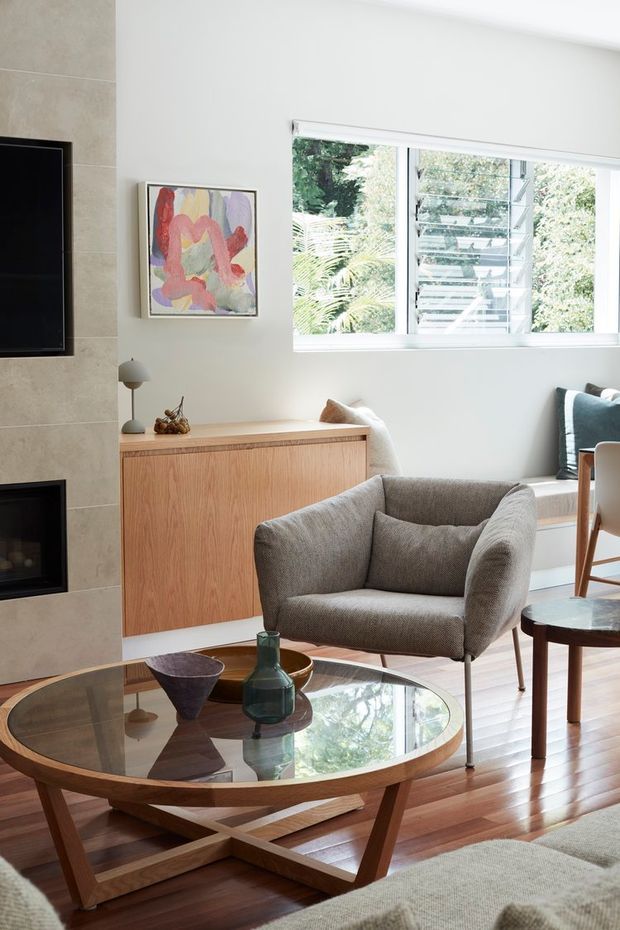 Bright, refined living room with mid-century-inspired furniture and warm timber flooring. The space features a large round glass-top coffee table with a timber geometric base, a sculptural upholstered armchair, a timber sideboard, a fireplace and television set into a pale stone feature wall, and a louvred window overlooking greenery. Neutral upholstery and timber finishes create a warm, airy atmosphere.