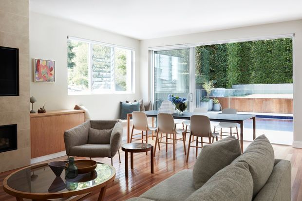 Bright open-plan living and dining area with a contemporary, minimalist fit-out. The room features warm timber flooring and built-in cabinetry, a large upholstered sofa and armchairs, a timber dining table with upholstered chairs, extensive glazing, and an adjoining outdoor terrace with a pool visible beyond. A fireplace and television wall are partially visible at the left.