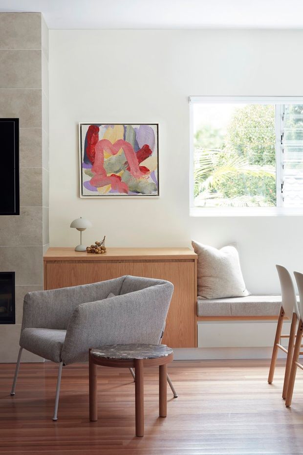 Bright contemporary living and dining area with warm timber flooring, a built-in timber sideboard and window seat, grey upholstered lounge chairs, a small round side table, abstract artwork, and a large louvred window. A tiled fireplace surround is visible at left.
