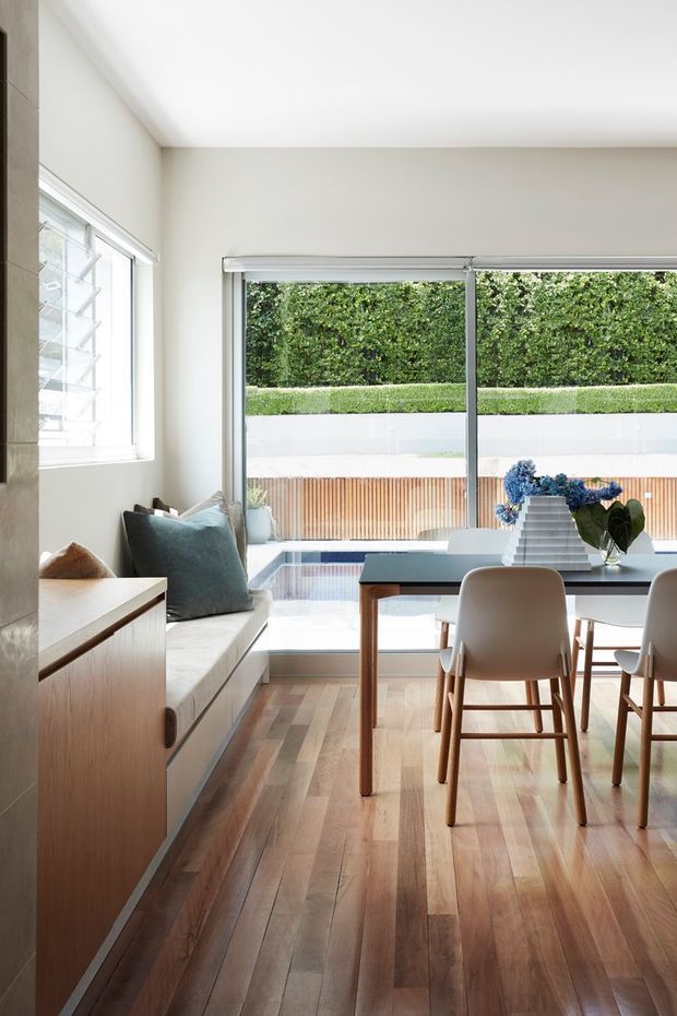 Bright contemporary dining area with a built-in window seat, timber dining table and upholstered chairs. The room has warm timber flooring, pale walls, large sliding glass doors and a side louvre window opening to a landscaped courtyard and pool-like water feature. Minimal decor includes blue cushions and flowers.