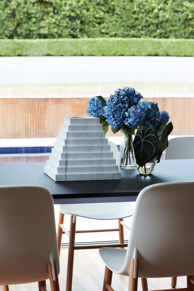 Bright contemporary dining area with a dark rectangular dining table, sculptural stepped stone centrepiece, and blue hydrangea arrangement in glass vases. Upholstered dining chairs with light timber legs surround the table. Large glazing and a timber privacy fence overlook a landscaped green hedge, creating an airy indoor-outdoor setting.