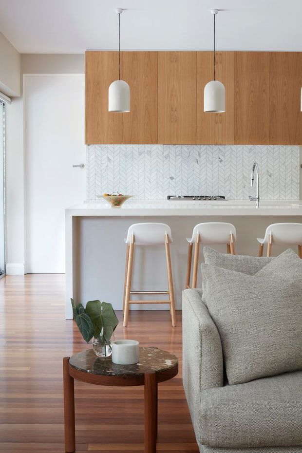 Bright open-plan kitchen and living area with warm timber cabinetry, a pale chevron-pattern tiled splashback, white stone benchtop, integrated breakfast bar, and three white pendant lights. The space features timber flooring, white bar stools, a light grey upholstered sofa, and a round dark timber coffee table with a stone top.