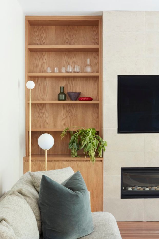 A close-up view of a refined living room featuring a full-height warm timber built-in shelving unit with open display shelves and lower cabinetry. Neutral stone-look wall tiles frame a wall-mounted television and linear fireplace, while a pale upholstered sofa, soft grey-green cushion, spherical lamps, decorative glassware, vases, and a leafy plant add warmth and texture.