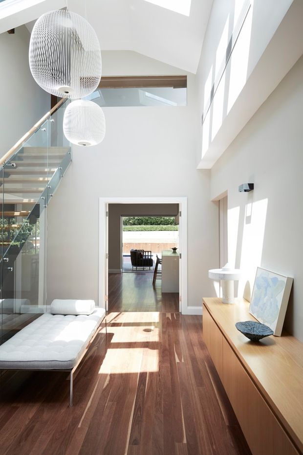 Bright, double-height contemporary hallway and living area with dark timber flooring, white walls, a glass-balustraded staircase, large pendant lights, clerestory-style glazing and a timber sideboard. A white daybed sits beside the staircase, while a connected room is visible through the doorway.