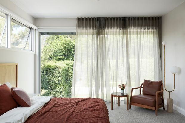 Bright, calm bedroom with large corner glazing, sheer full-height curtains, a low light-wood bedhead, and a rust-toned textured bedcover. A timber-framed leather lounge chair, small side table, and sculptural floor lamp sit beside the windows, with leafy greenery visible outside.