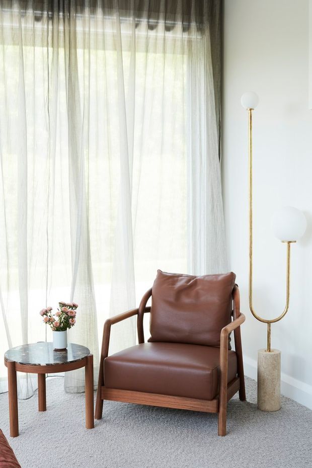 A bright, refined sitting corner featuring a mid-century-inspired brown leather armchair with timber arms and frame, a round timber side table with a dark stone-like top, sheer floor-to-ceiling curtains, and a sculptural brass floor lamp with globe shades. The room has pale walls and light grey carpet, creating a warm yet airy interior.