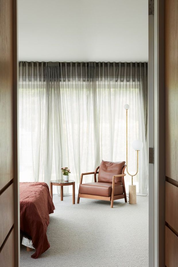 A calm, refined bedroom viewed through a doorway, featuring a low bed with rust-coloured bedding, a timber-framed leather lounge chair, a small round side table with flowers, sheer full-height curtains, and sculptural brass-and-globe floor lighting. Warm timber wall and door detailing frame the room, while pale carpet and abundant filtered daylight create a soft, airy atmosphere.