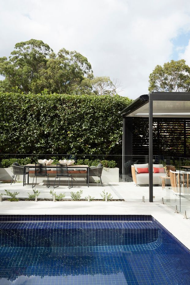 A contemporary outdoor entertaining area beside a deep blue tiled swimming pool. The space includes a long outdoor dining table with woven chairs, built-in planting, dense hedging, and a glass-enclosed pavilion or lounge with dark framing and louvred screening. The setting is bright and landscaped, with mature trees beyond the hedge.