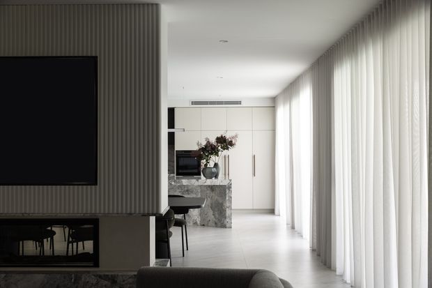 A refined open-plan contemporary interior viewed from a living area toward a compact kitchen and dining zone. Floor-to-ceiling sheer curtains line the right-hand wall, flooding the space with diffused natural light. A vertically ribbed feature wall with a wall-mounted television and dark fireplace anchors the foreground, while pale full-height cabinetry, a veined stone island, integrated appliances, and a small dark dining setting occupy the rear. The palette is restrained in whites, light greys, charcoal, and natural stone, creating a calm, minimalist atmosphere.
