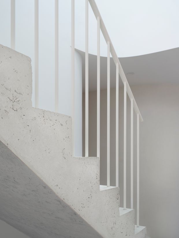 Minimalist interior detail of a concrete staircase with rough-textured pale concrete treads and side structure, paired with a slender white vertical metal balustrade. The composition is bright and monochromatic, emphasizing clean geometric lines and the stair's sculptural form.