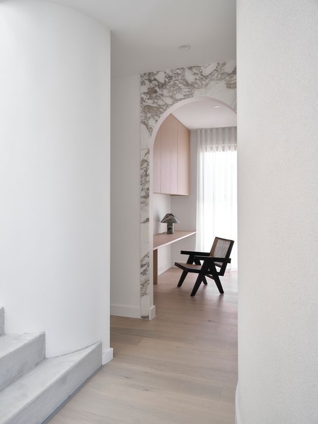A softly lit minimalist interior viewed from a hallway, looking through a rounded archway framed with dramatic marble-patterned surface into a compact study or seating nook. Pale timber flooring continues throughout, with a slim built-in desk and blush-pink cabinetry beside a tall sheer-curtained window. A dark timber and woven lounge chair sits in the nook, while curved white walls and a concrete stair edge are visible in the foreground.