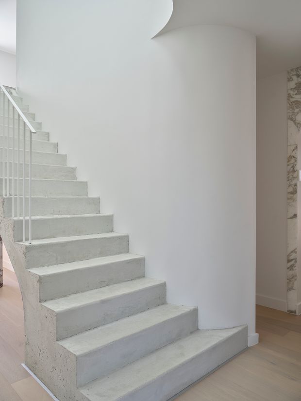 Minimalist interior stairway with a sculptural curved white wall and broad monolithic pale concrete steps. A slim white metal balustrade runs along the open side, while light timber flooring and a marble-patterned wall surface are visible beyond the stair. The space is bright, restrained, and contemporary.