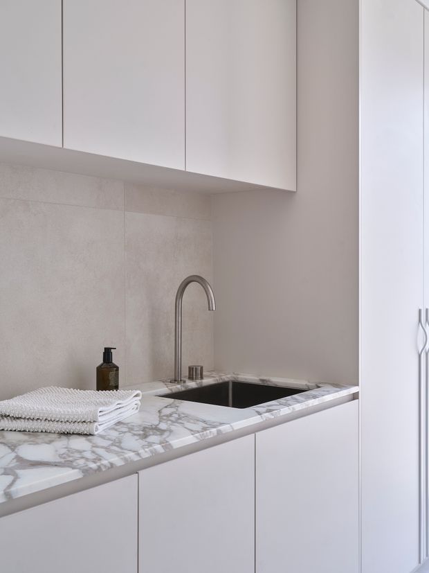 A compact, refined laundry or utility space with seamless white flat-panel cabinetry, a pale stone-tile splashback, and a veined marble benchtop incorporating a deep undermount sink. A tall curved metal tap, folded white towels, and a soap dispenser sit on the counter. The space is softly lit with a restrained neutral palette and minimal detailing.