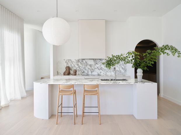 Bright, minimalist contemporary kitchen with a large white marble island and matching marble splashback. The island has softly rounded ends, integrated cabinetry, a sink and tap, and two light timber bar stools. White walls, sheer full-height curtains, pale timber flooring, a large spherical pendant, and an arched doorway create an airy, refined interior. Greenery and ceramic vessels provide subtle natural accents.