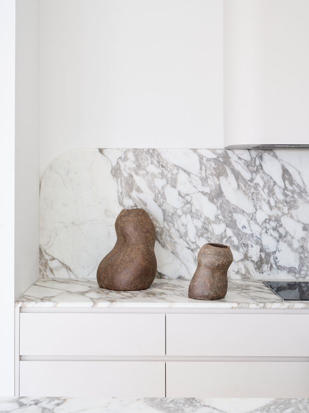 Close-up of a refined white built-in kitchen or living-area joinery composition with a dramatic veined marble backsplash and matching benchtop, flat-front white drawers, and two sculptural brown decorative vessels. The image is bright, minimalist, and detail-focused, with a partial dark cooktop or appliance visible at the right edge.