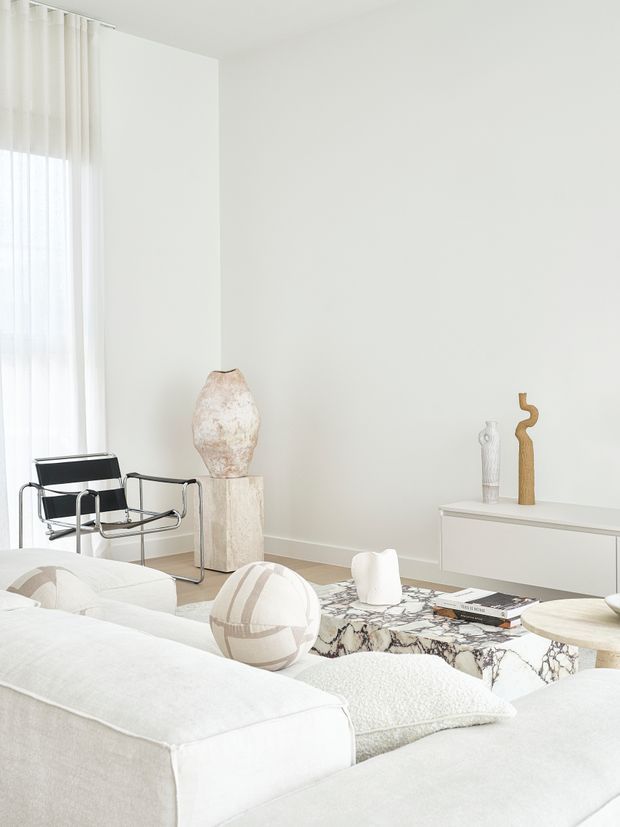 Bright, minimalist living room with a predominantly white palette. A large white sectional sofa occupies the foreground, facing a low marble-patterned coffee table with books and sculptural ceramics. A black leather-and-chrome lounge chair sits beside a tall window dressed in sheer white curtains, while a pale stone pedestal supports a large organic ceramic vase. A streamlined white sideboard with sculptural objects completes the room. The space has a refined, airy contemporary feel with abundant natural light.