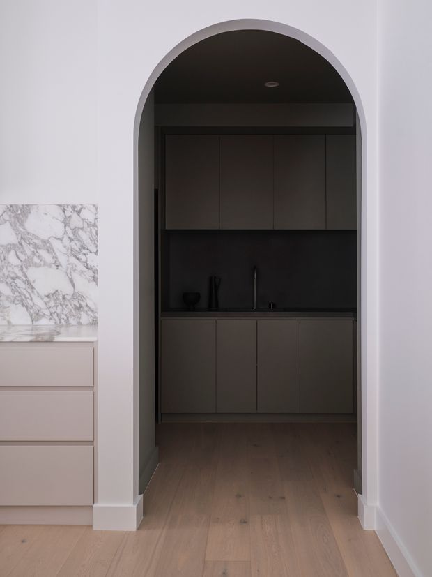 A refined interior view through a tall arched opening into a compact, dark-toned kitchen or scullery. The space features floor-to-ceiling matte dark cabinetry, a dark benchtop and splashback, a black sink and tapware, and warm timber flooring. At left, a pale pink-toned cabinet run is topped with a dramatic white marble surface with grey veining. The surrounding walls and arch are white, creating strong contrast with the moody cabinetry.