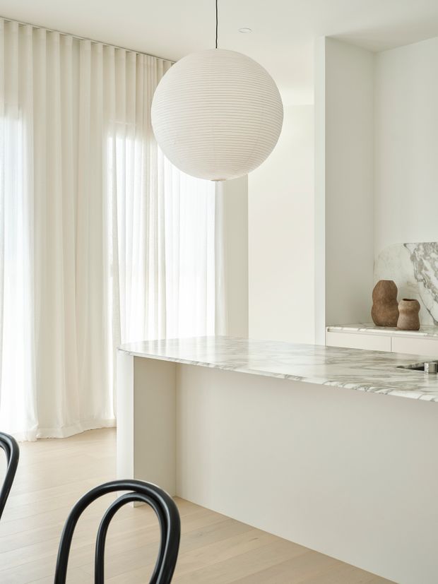 Bright, minimalist kitchen interior with a long marble island or peninsula, pale timber flooring, sheer full-height curtains, white walls and cabinetry, and a large ribbed spherical pendant light. Black curved dining chairs and a small display niche with sculptural vessels add contrast and warmth.