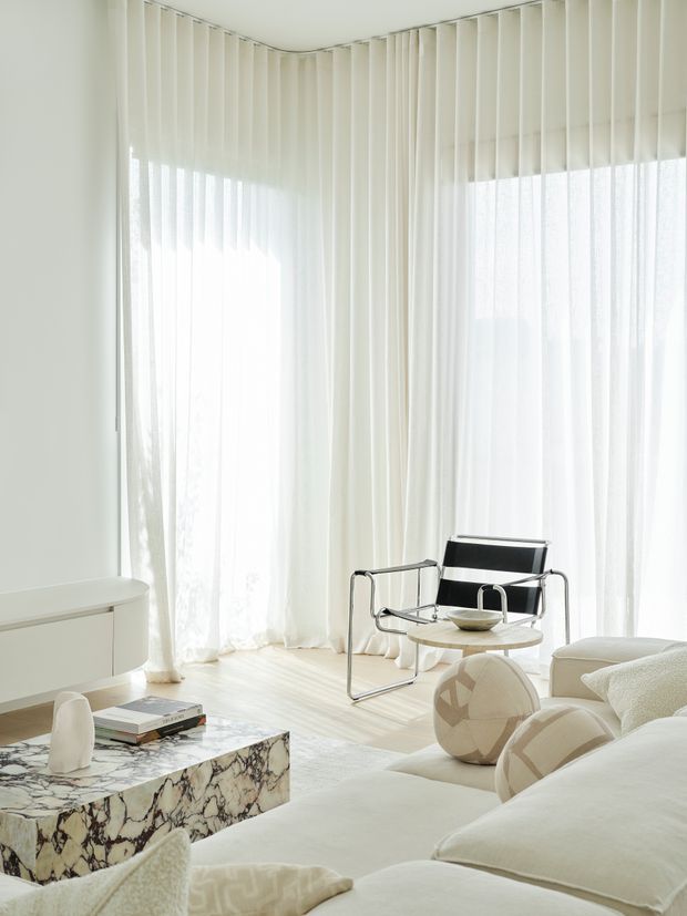 Bright, airy living room with floor-to-ceiling sheer curtains wrapping around corner glazing. The space features a white sofa, black leather-and-chrome lounge chair, small round coffee table, sculptural neutral cushions, a low white sideboard, and a striking veined stone coffee table. The overall composition is refined and minimalist with abundant natural light.