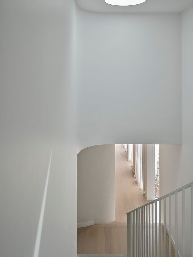 A bright minimalist interior stairway with white plaster walls, a curved opening, pale timber flooring, and a simple grey metal balustrade. The stair looks toward a light-filled corridor lined with tall windows and sheer curtains. A recessed circular ceiling light adds a clean contemporary detail.