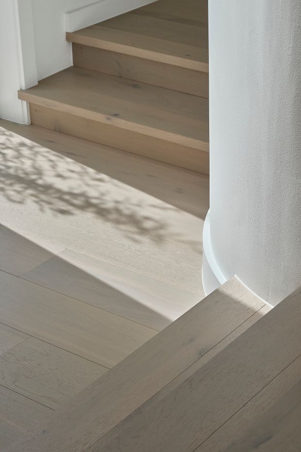 Close-up interior view of a light natural timber staircase and adjoining timber flooring, with simple white plastered walls and soft sunlight casting leafy shadows across the floor. The image focuses on the stair construction, pale wood tones, and clean minimalist detailing.