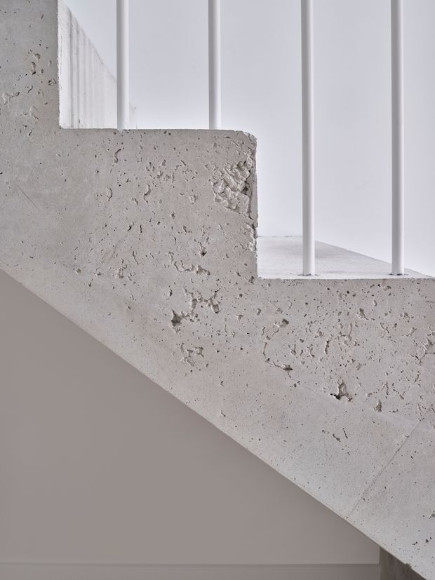 Close-up architectural detail of a raw, textured concrete staircase or stair landing with a diagonal underside and slim white metal balustrade. The surrounding walls and vertical panelling are white, creating a restrained minimalist composition focused on the contrast between rough concrete and smooth painted surfaces.