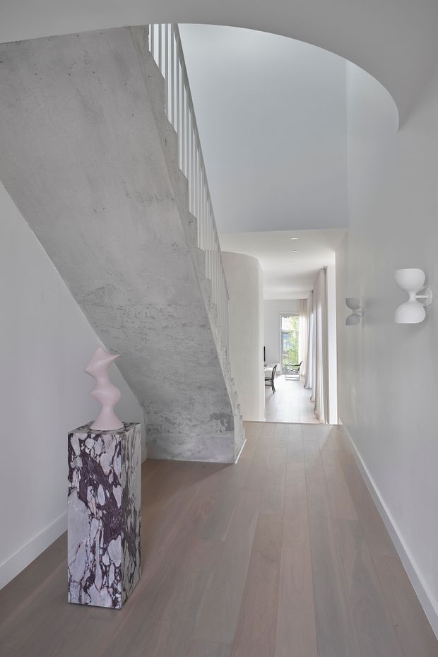 A bright minimalist hallway with a sculptural exposed concrete staircase and slim white balustrade. Pale timber flooring runs toward a glazed opening at the far end, while a dramatic veined stone pedestal with a pink sculptural object and simple white wall sconces add visual interest. The space has clean white walls, curved ceiling detailing, and strong natural light.