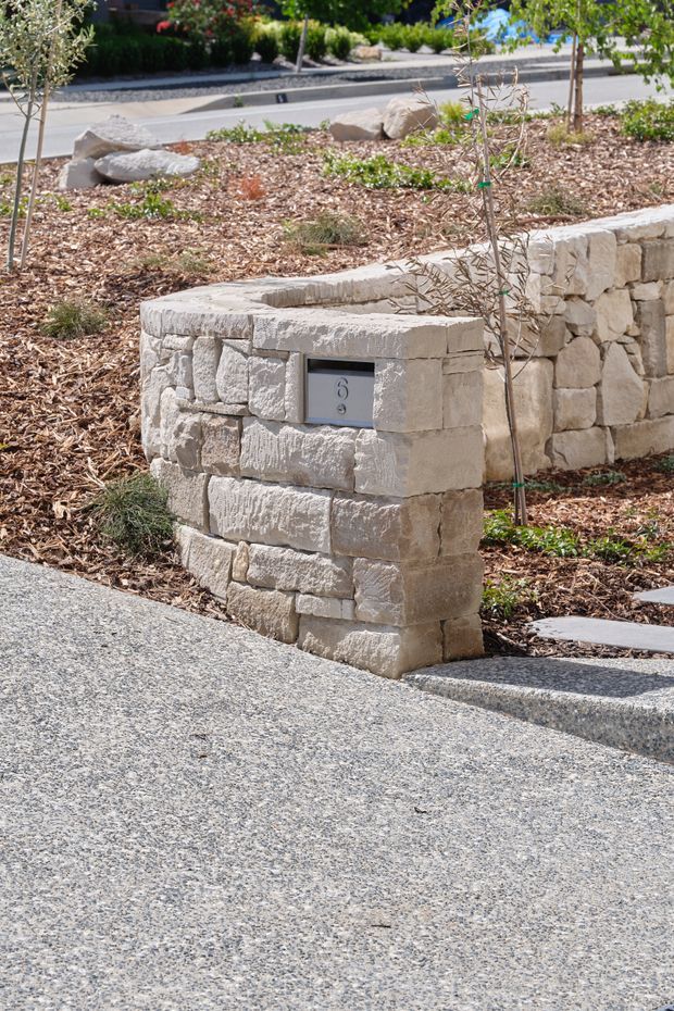 A landscaped residential streetscape featuring a curved retaining wall and square pillar constructed from rough-cut light beige stone blocks. The pillar incorporates a small metallic house-number plaque, while young trees, mulch beds, planted landscaping, stepping pavers, and a textured aggregate driveway surround the masonry. The scene is bright and natural, with a neat contemporary garden design.