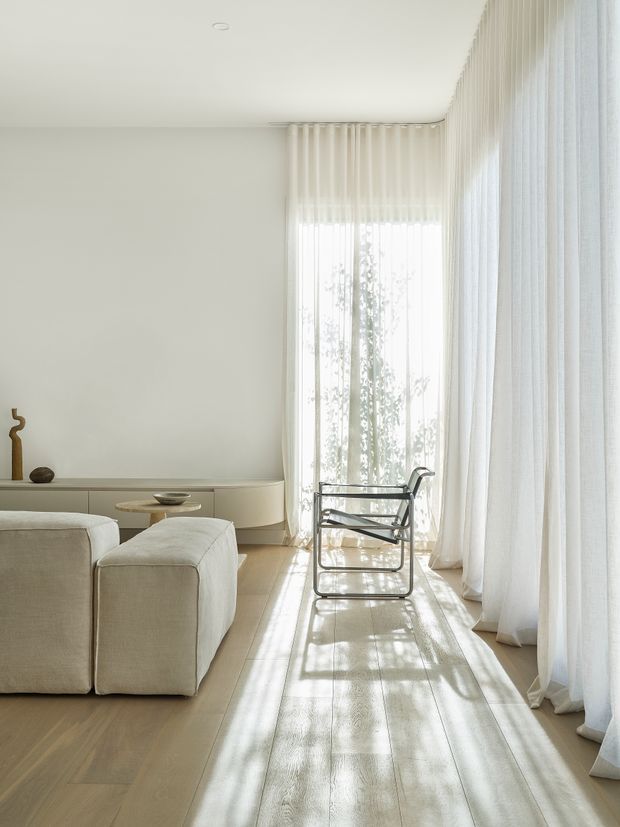 A bright, minimalist living room with floor-to-ceiling sheer white curtains, pale timber flooring, a low curved built-in media console, and a cream upholstered sofa. A black-framed lounge chair sits beside the tall window, while sunlight creates strong linear shadows across the floor. Neutral finishes, sparse decor, and soft natural light create an airy, serene atmosphere.