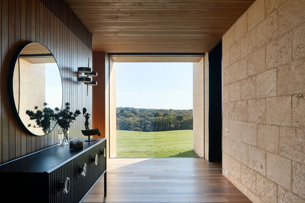 A refined modern entry hall with a dark vertically panelled built-in console and oval mirror on the left, a textured pale stone block wall on the right, and a timber-lined ceiling. Full-height glazing at the end frames an expansive green lawn and wooded landscape, while warm timber flooring and sculptural lighting create a calm, contemporary atmosphere.