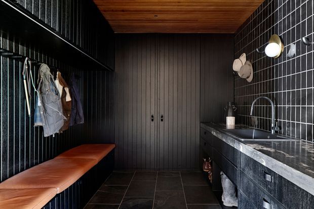 A dark, compact mudroom or entry storage space with black vertical wall panelling, a built-in hanging rail and open shelf on the left, a long tan leather bench, central black storage doors, and a dark stone-topped utility counter with sink on the right. Black tiled walls and floor contrast with a warm timber-lined ceiling and brass-toned wall lighting.