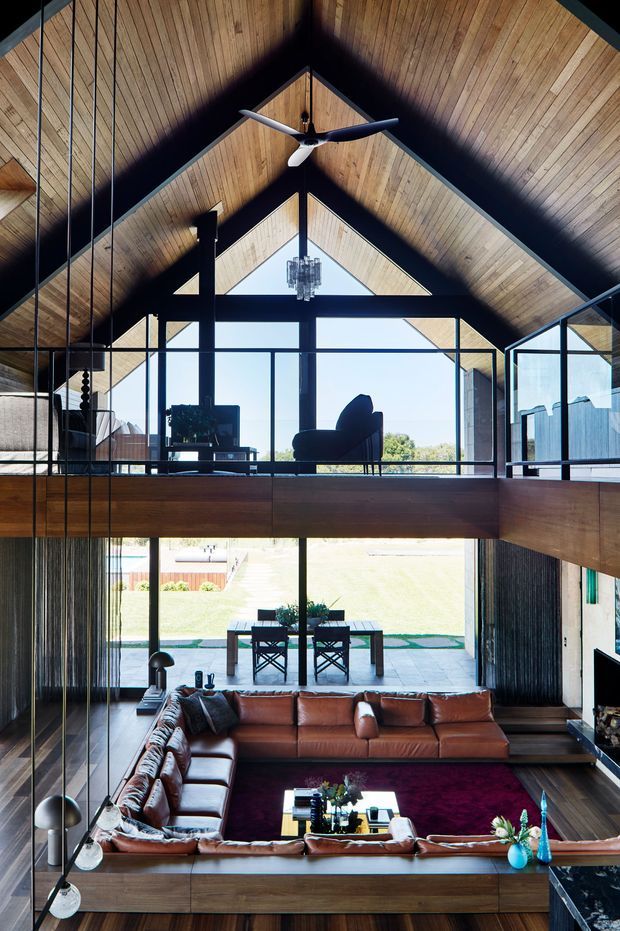 A dramatic double-height living room with a mezzanine level, exposed timber gabled ceiling, dark structural beams, extensive floor-to-ceiling glazing, and a large suspended ceiling fan. The lower level contains a brown leather sectional around a dark rug and coffee table, while the upper level has a lounge chair and chandelier. The glazing opens visually toward a landscaped outdoor area and dining terrace.