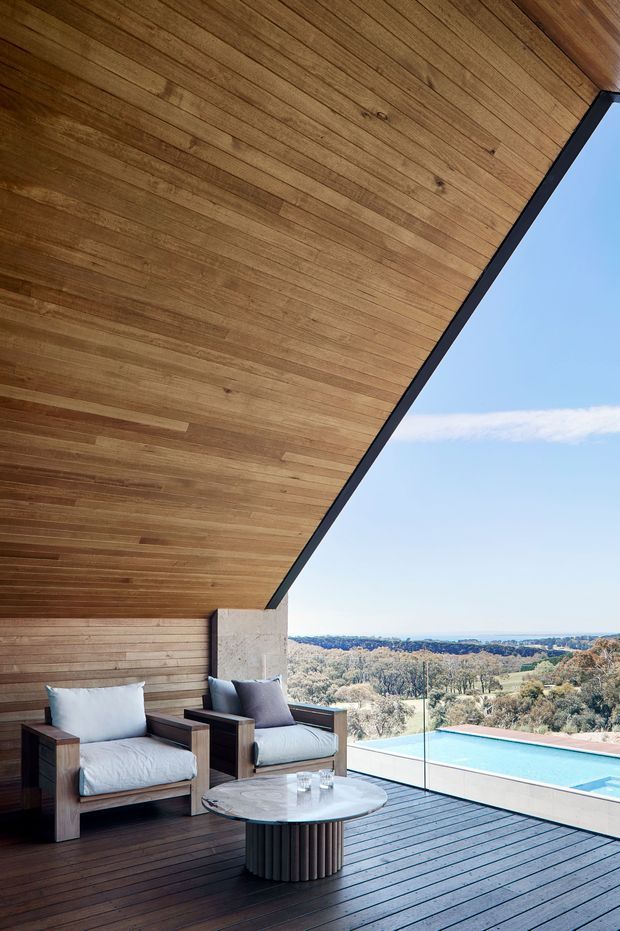 A covered outdoor terrace with a dramatic sloping timber-lined ceiling and dark timber decking. Two cushioned timber-framed lounge chairs and a round coffee table sit beside frameless glazing, overlooking a swimming pool and expansive treed landscape. The structure has a strong contemporary pavilion-like form with a prominent angled roof plane.