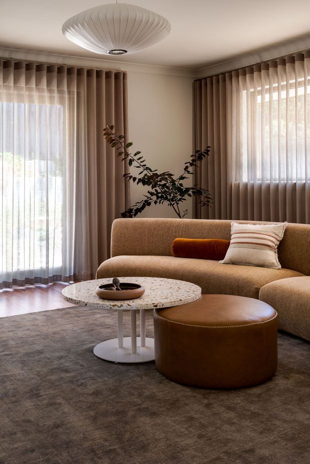 A warm, refined living room with a curved tan upholstered sofa, large rounded leather ottoman, terrazzo-style round coffee table, layered sheer and full-height taupe curtains, timber flooring, and a sculptural pleated ceiling pendant. A branch arrangement adds an organic accent, while soft natural light creates a calm, contemporary atmosphere.