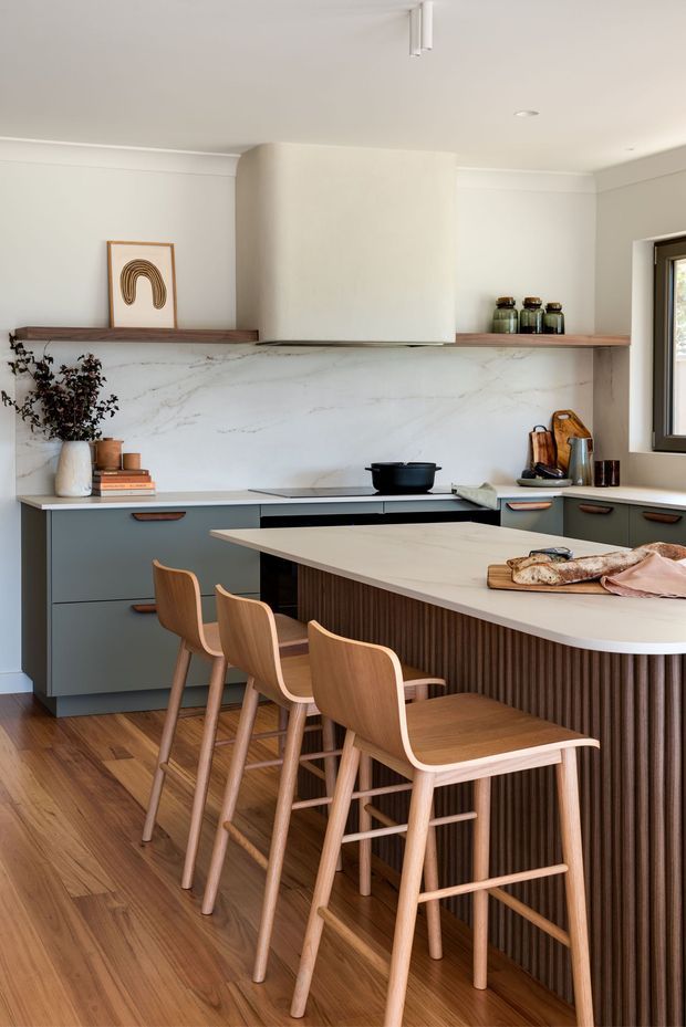 A warm, contemporary kitchen with a large island and integrated seating. The space features muted grey-green cabinetry, a pale stone waterfall benchtop, vertically fluted timber island cladding, a veined stone splashback, timber flooring, and light timber bar stools. A long timber shelf spans the splashback wall, with understated ceramic and glass accessories. Soft neutral walls, streamlined cabinetry, and natural timber create a refined, welcoming interior.