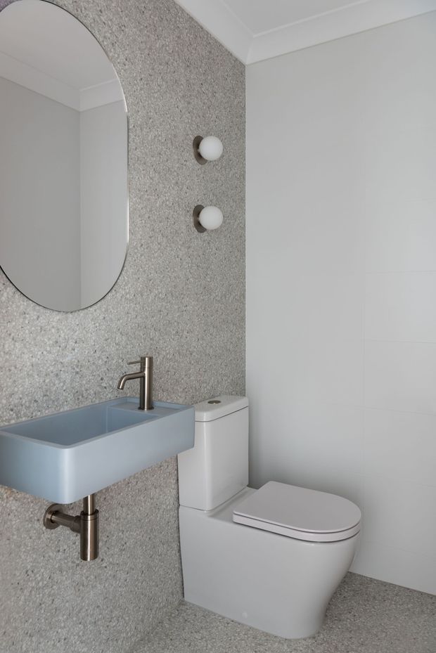 A compact contemporary bathroom featuring a wall-mounted pale blue-grey basin with brushed metal tap and exposed plumbing, a white toilet, and a large rounded-rectangle mirror. The walls and floor are finished in speckled grey terrazzo-like material, with white tiled wall surfaces, simple globe wall lights, and white decorative ceiling moulding.