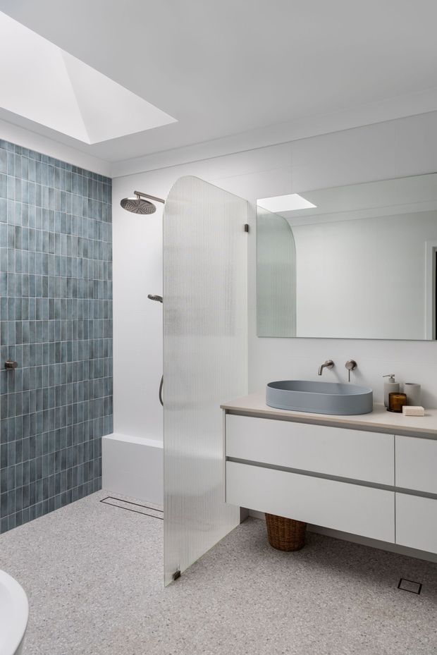 Bright contemporary bathroom with a walk-in shower, blue-grey vertical wall tiles, terrazzo-style flooring, a fluted translucent glass shower screen, wall-mounted white vanity with a grey vessel basin, large mirror, skylight, and brushed metal fixtures.
