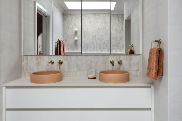 A contemporary double-vanity bathroom with two round terracotta vessel sinks on a pale stone-look benchtop, a wide mirrored wall cabinet, warm brass wall-mounted taps, pale textured wall tiles, and a white floating vanity. A skylight or recessed ceiling light provides bright illumination, while rust-coloured towels add warmth.