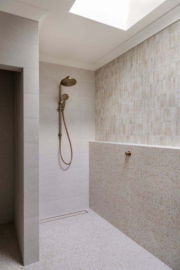 A contemporary walk-in shower bathroom with a frameless open entry, large pale wall tiles, a textured vertical feature-tile wall, and matching terrazzo-like floor and half-height partition. The shower fittings are warm brass, and a large skylight provides soft natural illumination.